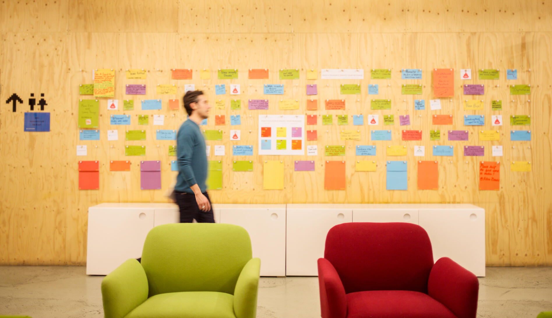 Man walking pass wall of post-it notes and sketches on a plywood wall with sofas in foreground.